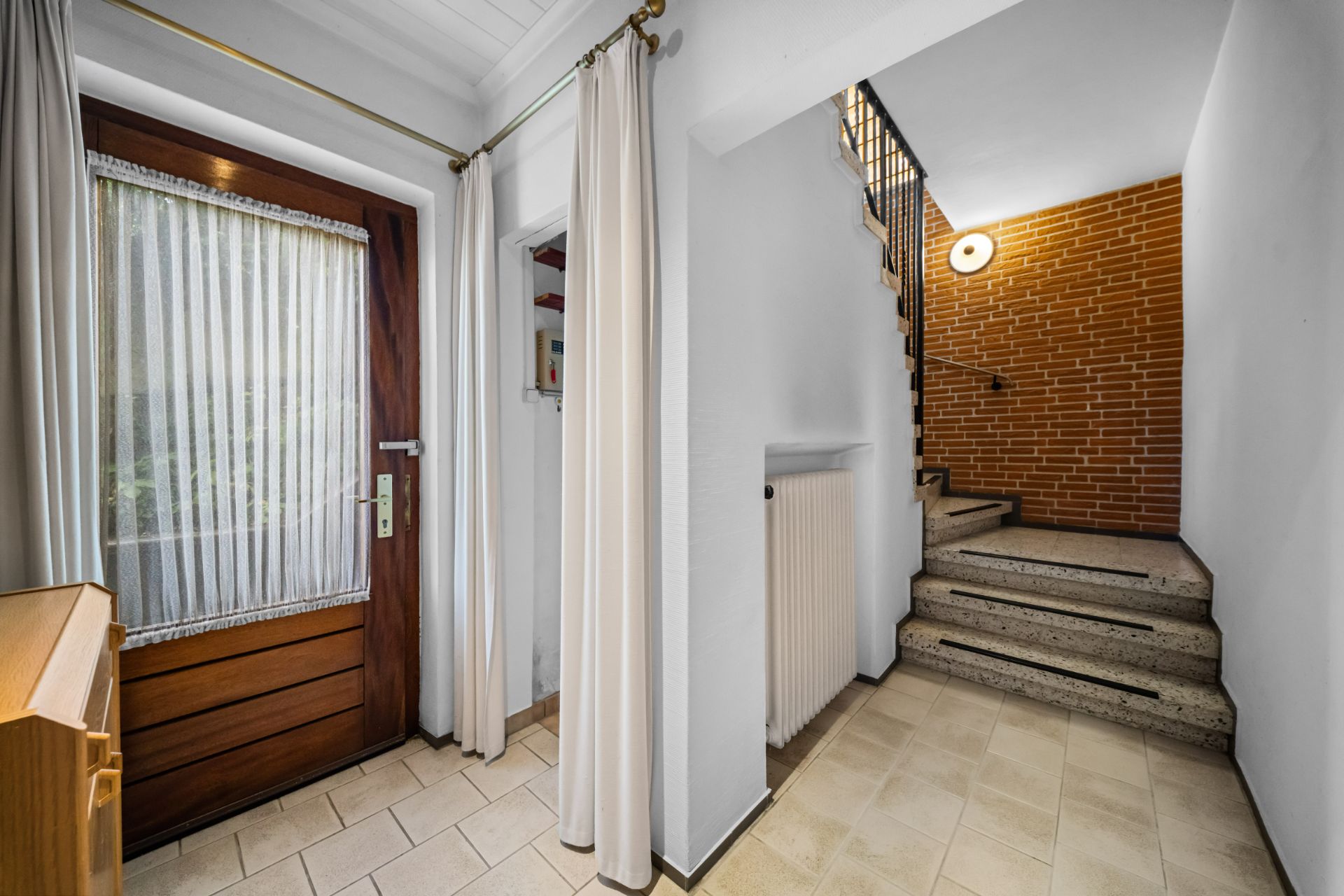 Eingangsbereich mit Holztür, Vorhängen, Heizung und Treppe neben Ziegelwand.