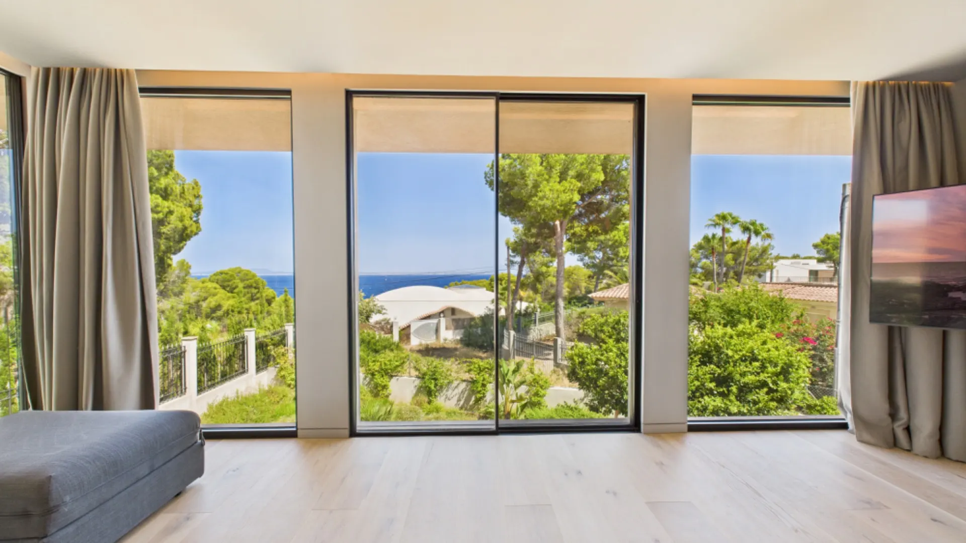 Helles Wohnzimmer mit großen Fenstern und Blick auf Garten, Bäume und das Meer.
