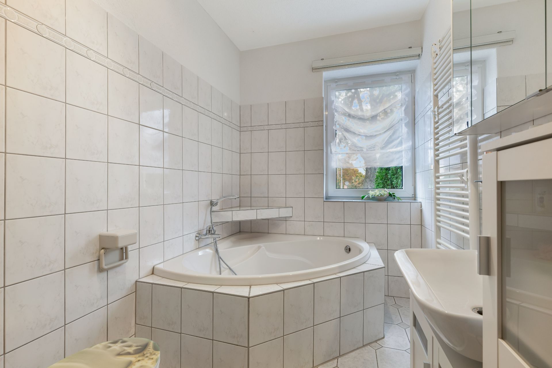 Helles Badezimmer mit Eckbadewanne, Waschbecken und Fenster mit Vorhang.
