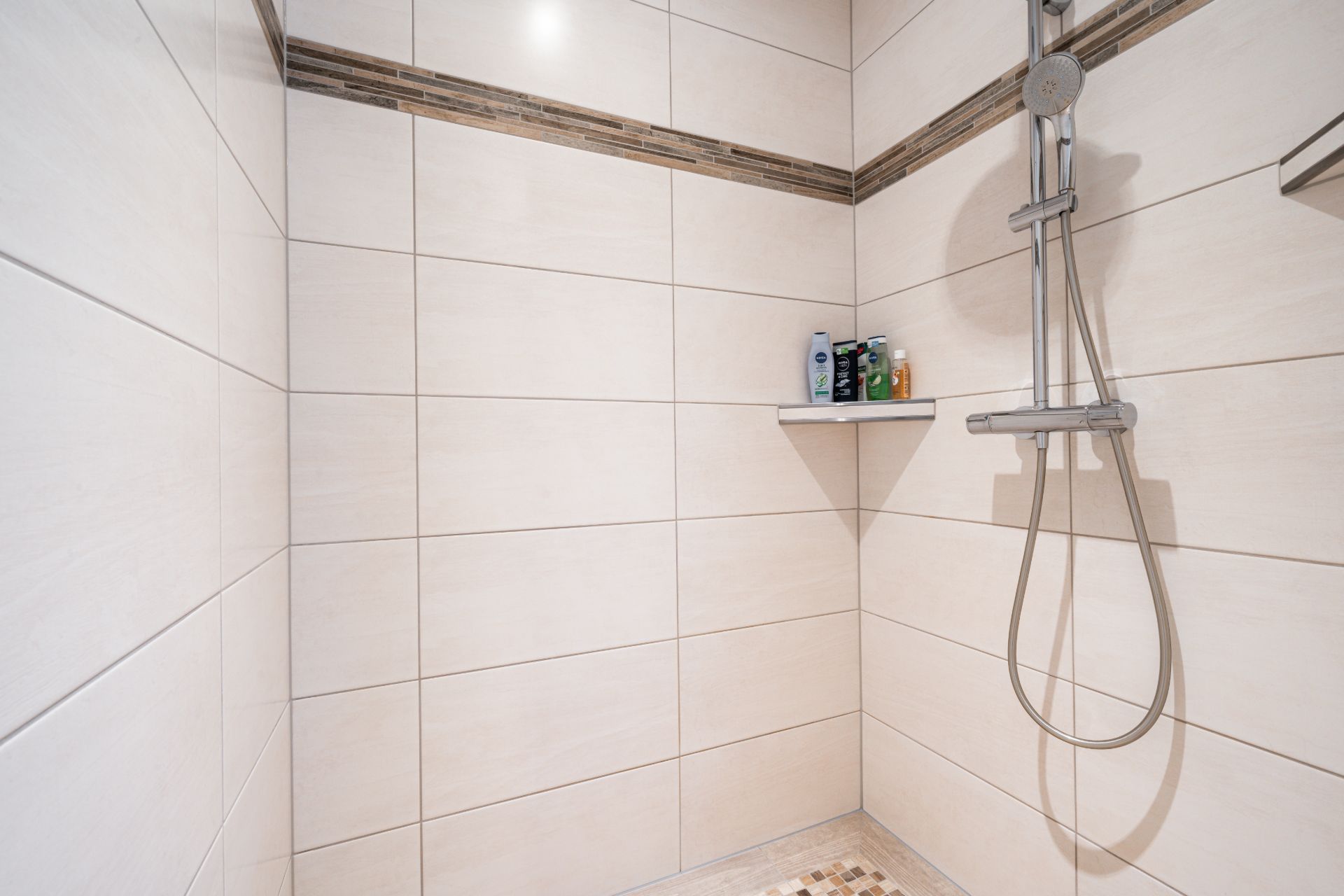 Dusche mit hellen Fliesen, Regendusche und Ablage mit Pflegeprodukten.