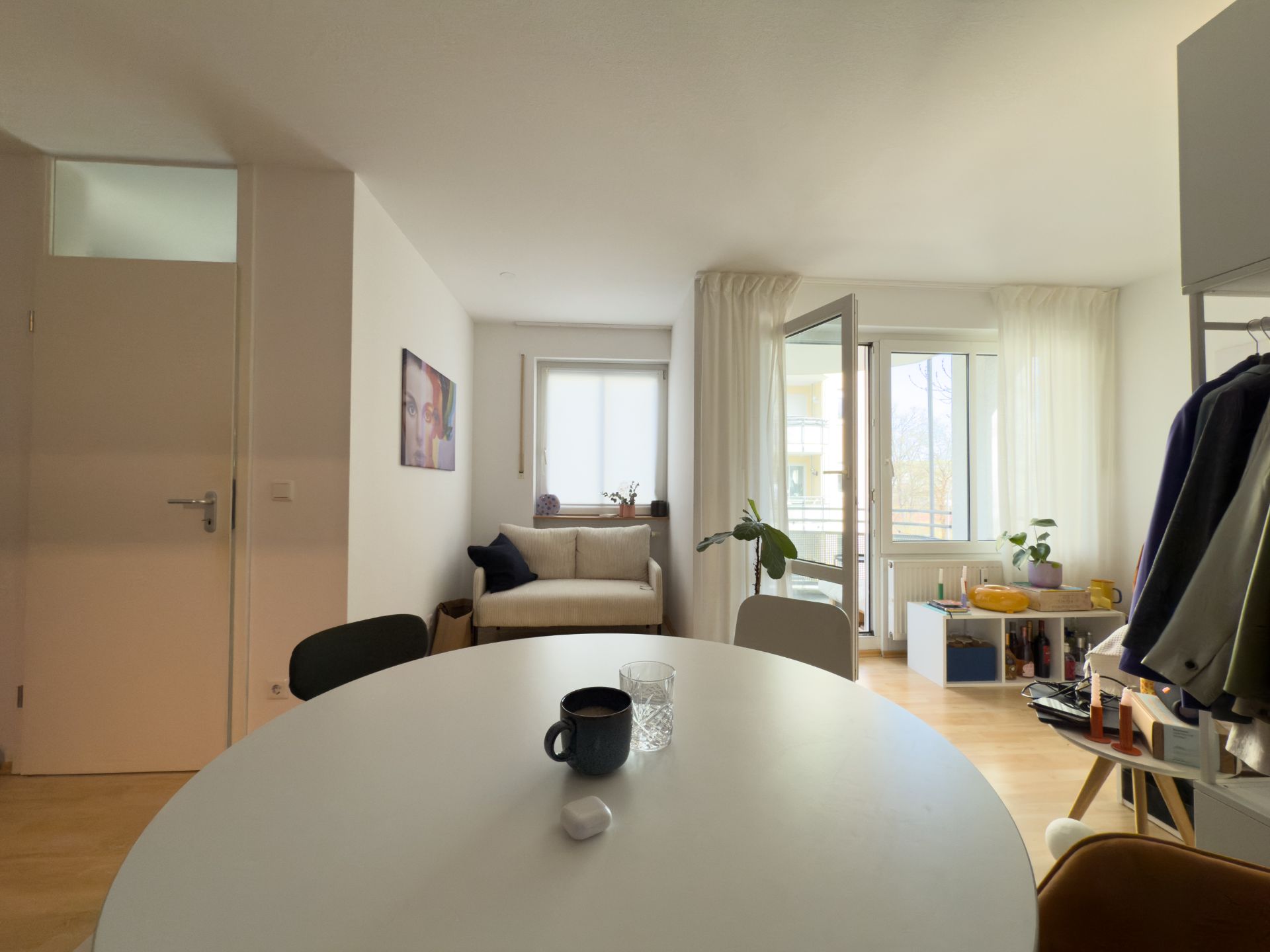 Helles Wohnzimmer mit Sofa, Esstisch, offener Tür zum Balkon und Pflanzen am Fenster.