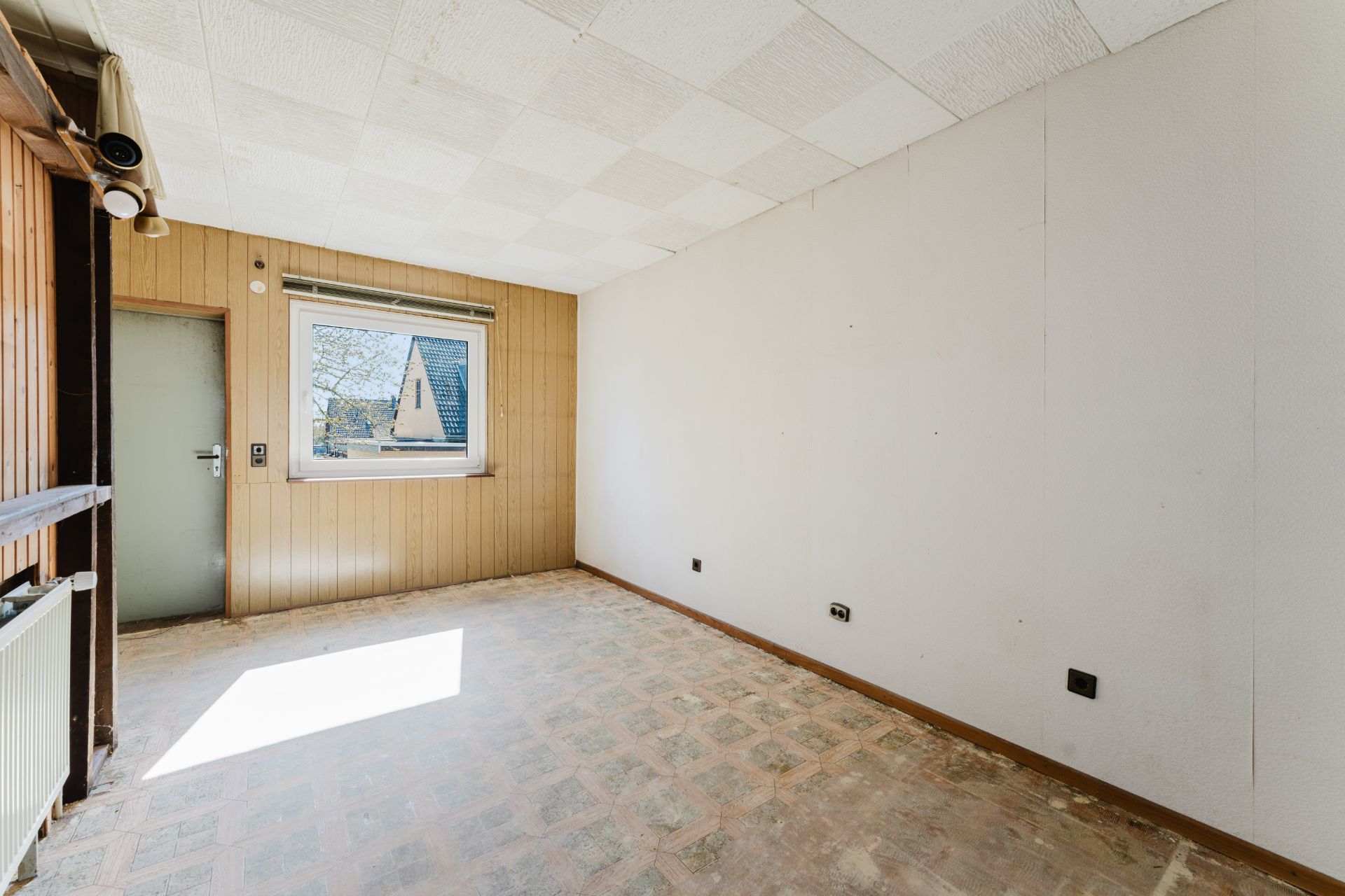 Leerer Raum mit Fenster, Holzwand und unbelegtem Boden, Tür links neben dem Fenster.