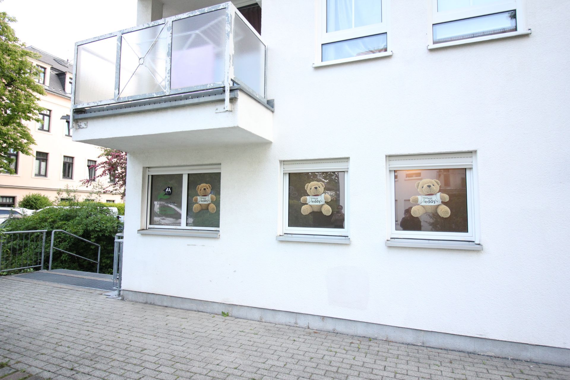 Zwei Fenster mit Teddybären und Namensschildern in einer weißen Wohnhausfassade.