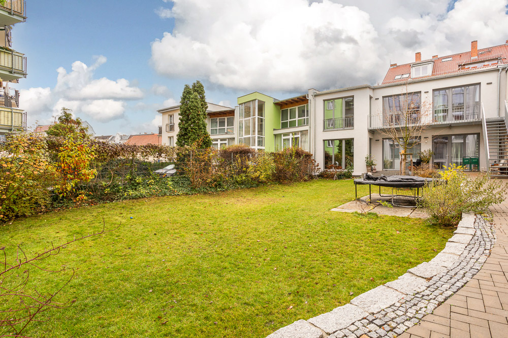 Grüner Garten mit Rasenfläche, kleinem Baum, Trampolin und Wohngebäuden im Hintergrund.