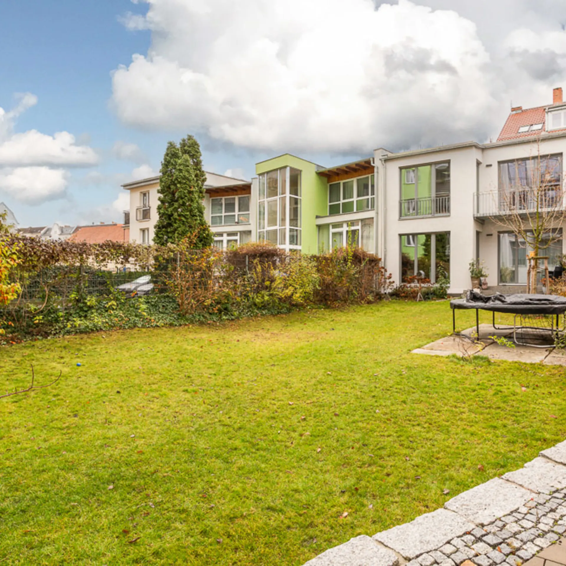 Grüner Garten mit Rasenfläche, kleinem Baum, Trampolin und Wohngebäuden im Hintergrund.
