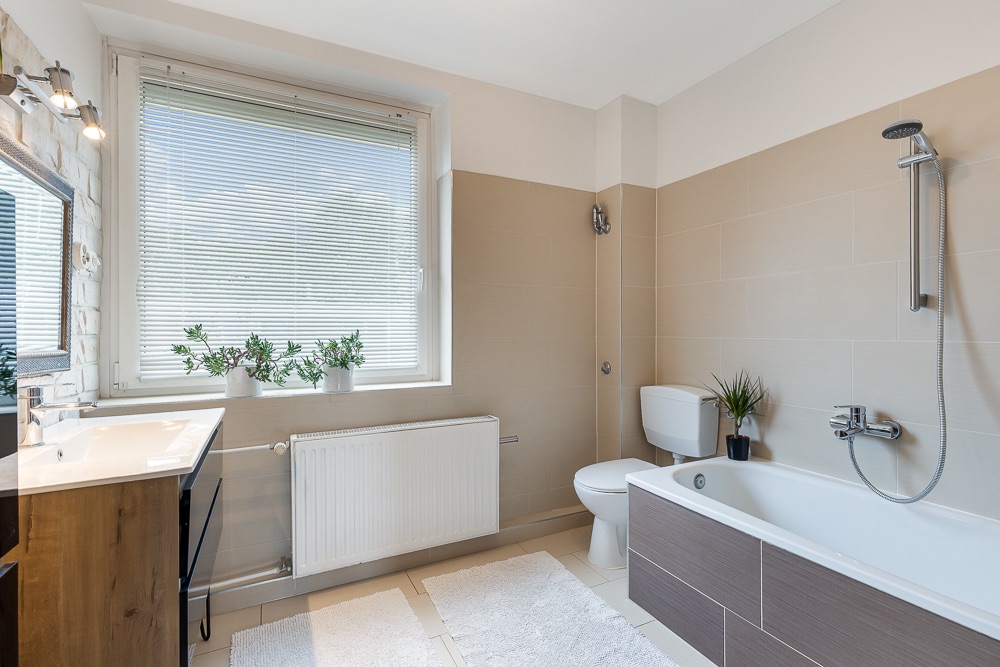 Helles Badezimmer mit Badewanne, Toilette, Waschbecken, großem Fenster und Pflanzen auf der Fensterbank.
