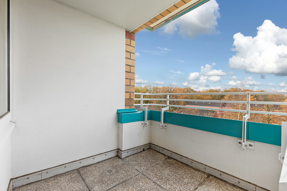 Balkon mit weißen Wänden, türkisfarbenen Geländern und Ausblick auf Bäume unter blauem Himmel.