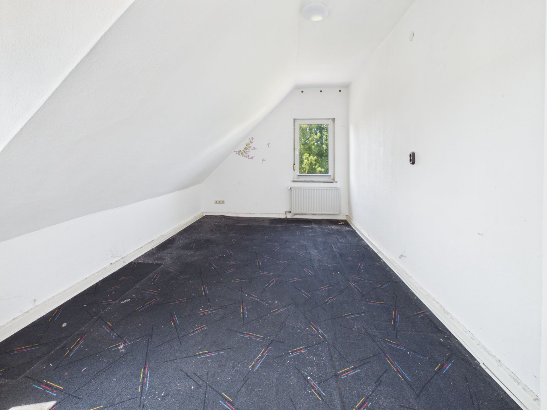Leeres Zimmer mit schräger Decke, dunklem Teppichboden und kleinem Fenster mit Blick ins Grüne.
