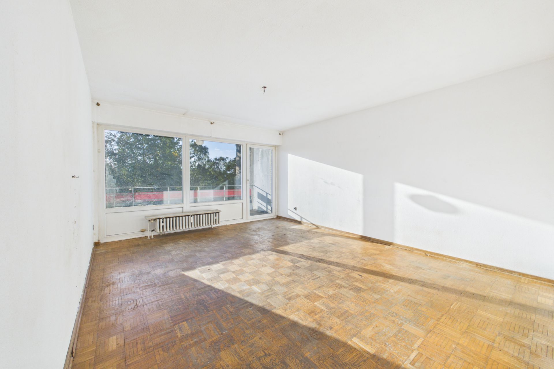 Leerer Raum mit Parkettboden, großen Fenstern und Heizkörper unter dem Fenster.