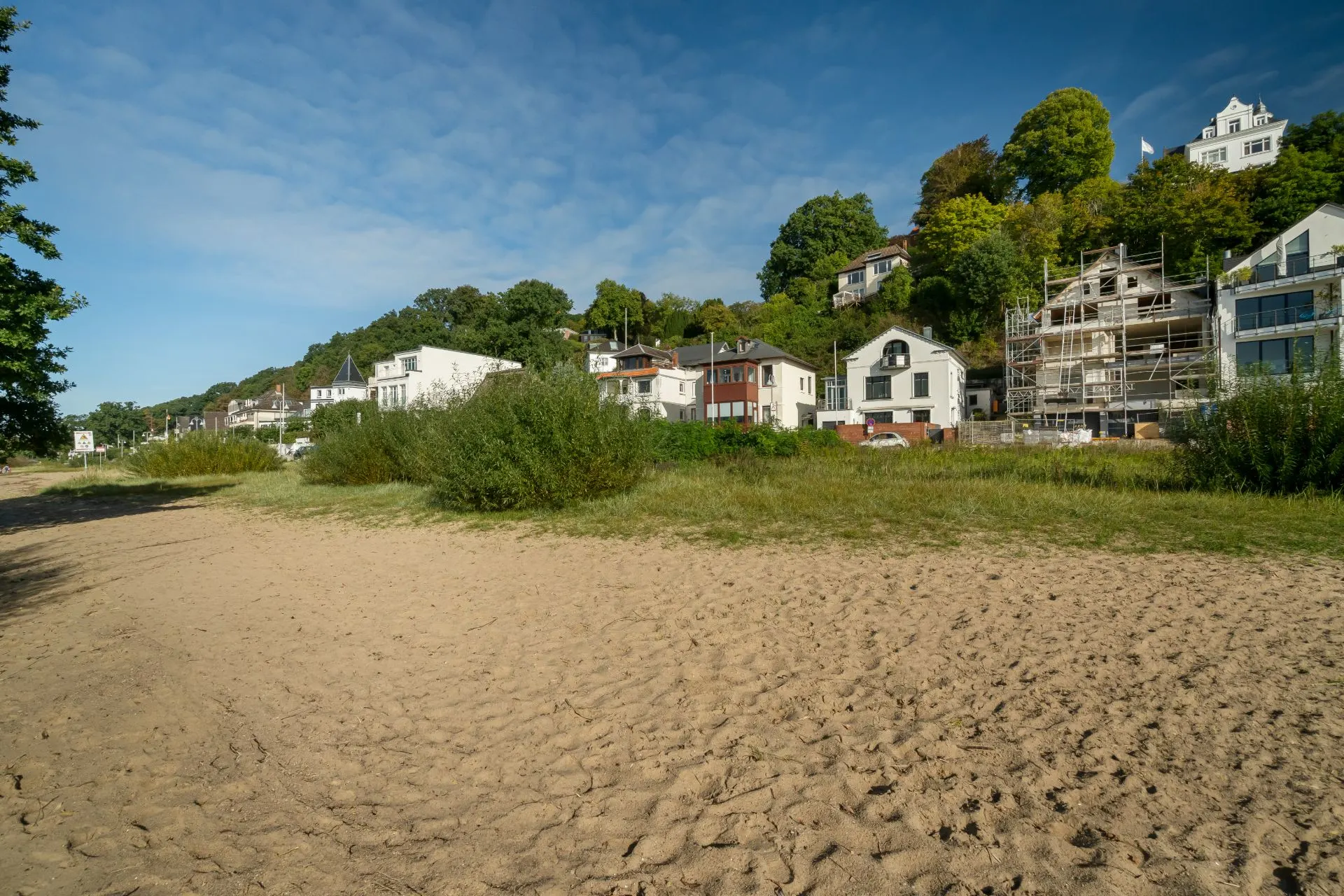 Mehrere Häuser stehen am Hang hinter einem Sandstrand, einige von Bäumen umgeben, eines im Bau.