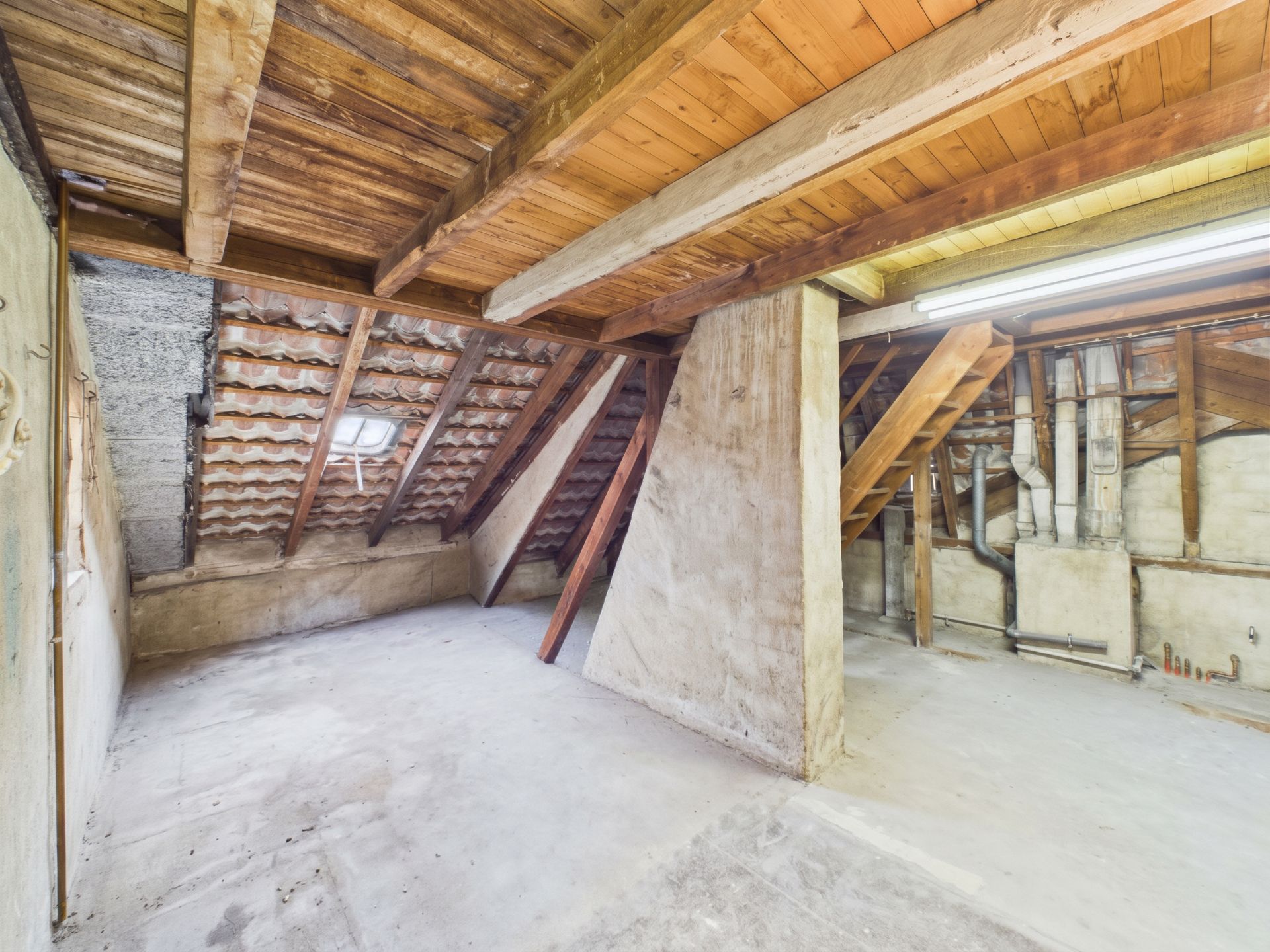 Leerer Dachboden mit sichtbaren Holzbalken, Treppe und kleinem Fenster.