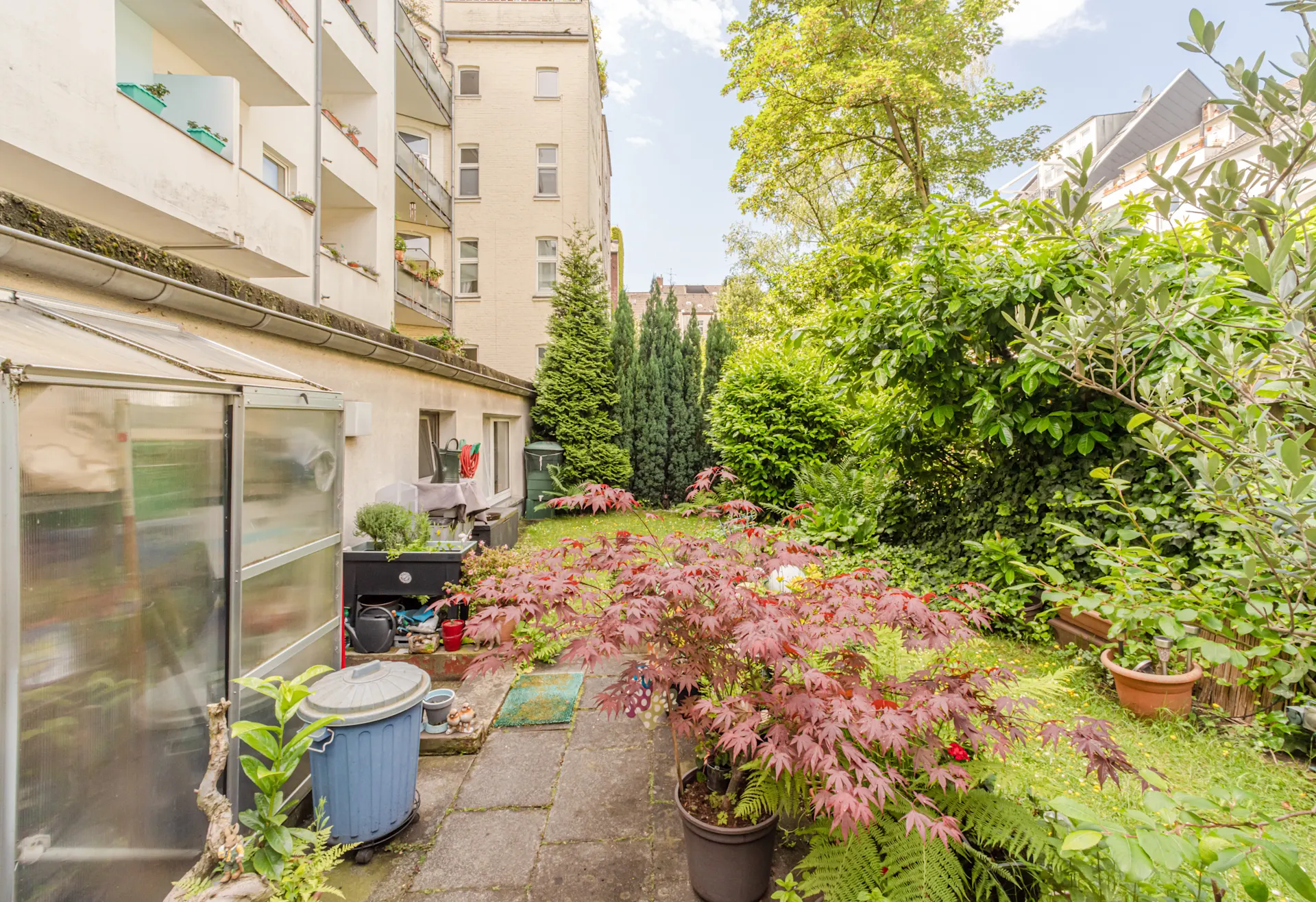 Begrünter Garten mit Pflanzen, Bäumen, Terrasse und einem kleinen Gewächshaus an einem Wohnhaus.