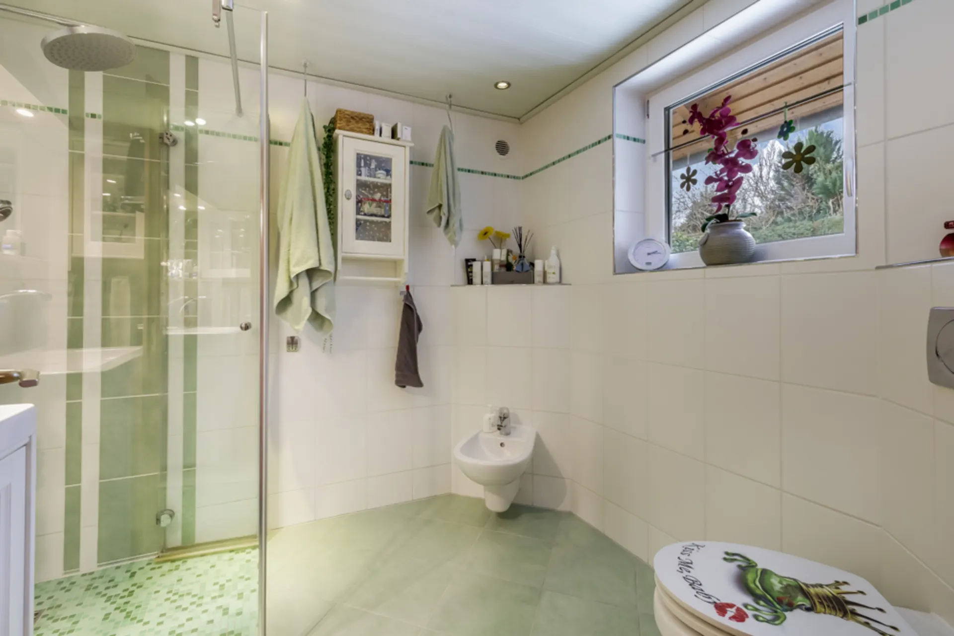 Modernes Badezimmer mit Dusche, Bidet, Fenster und Dekoration, grüne Fliesen am Boden.