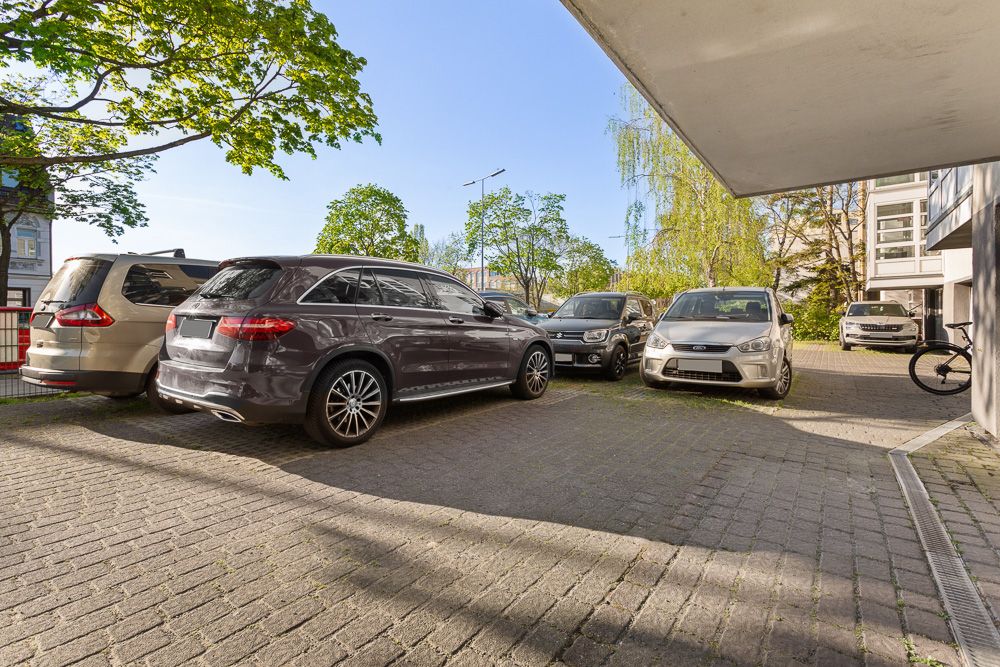 Mehrere Autos parken auf einem gepflasterten Parkplatz neben einem Wohnhaus, umgeben von Bäumen.
