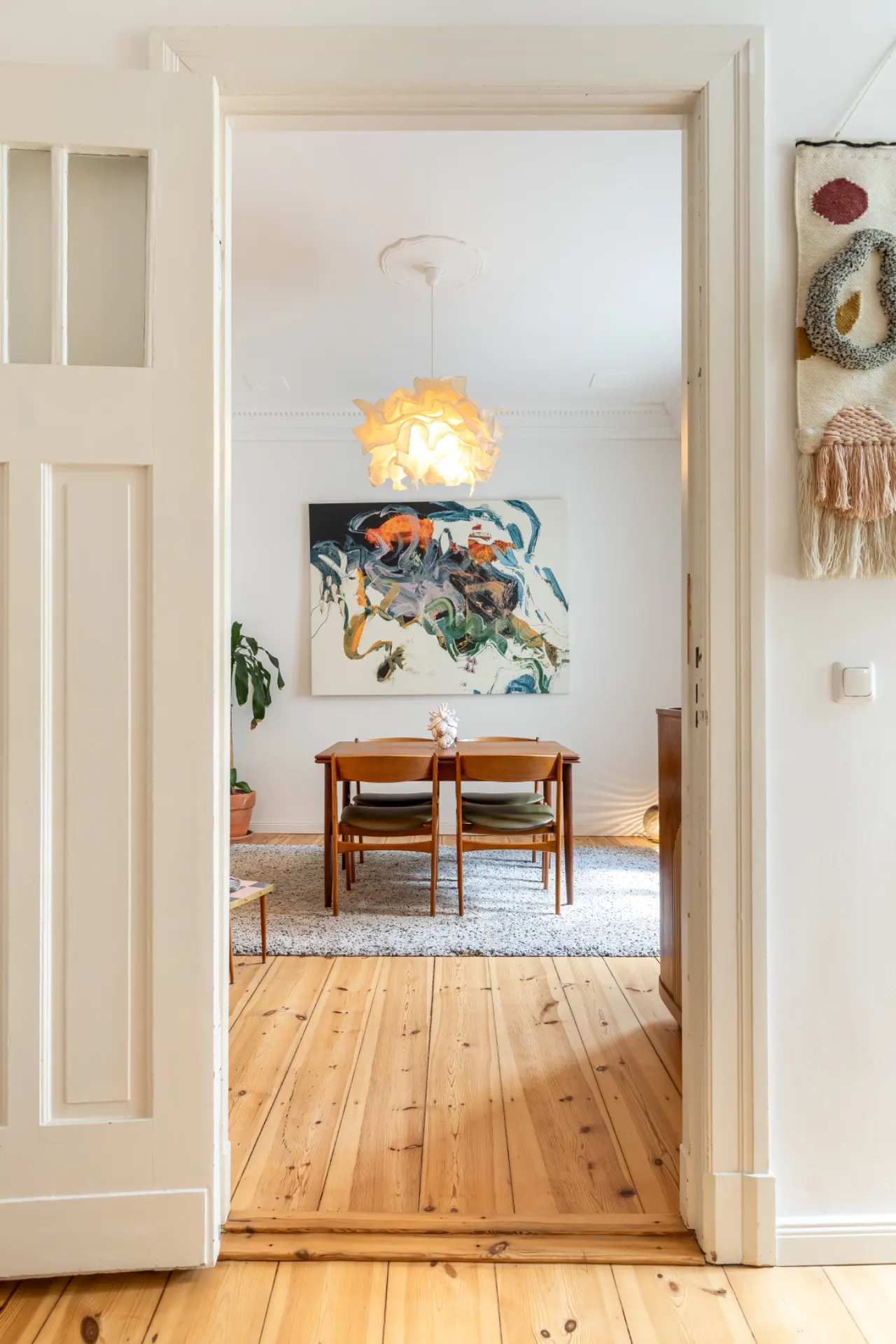 Esszimmer mit Holzboden, vier Stühlen, Kunstwerk an der Wand und Deckenlampe.