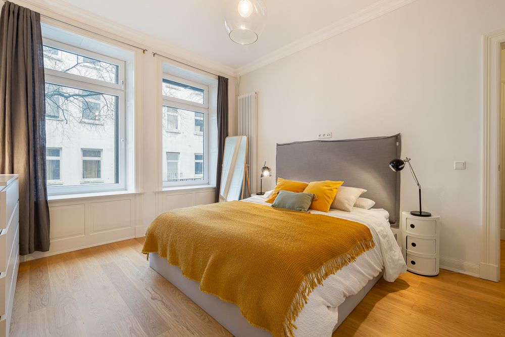 Helles Schlafzimmer mit Doppelbett, gelber Decke, Nachttisch und großen Fenstern.