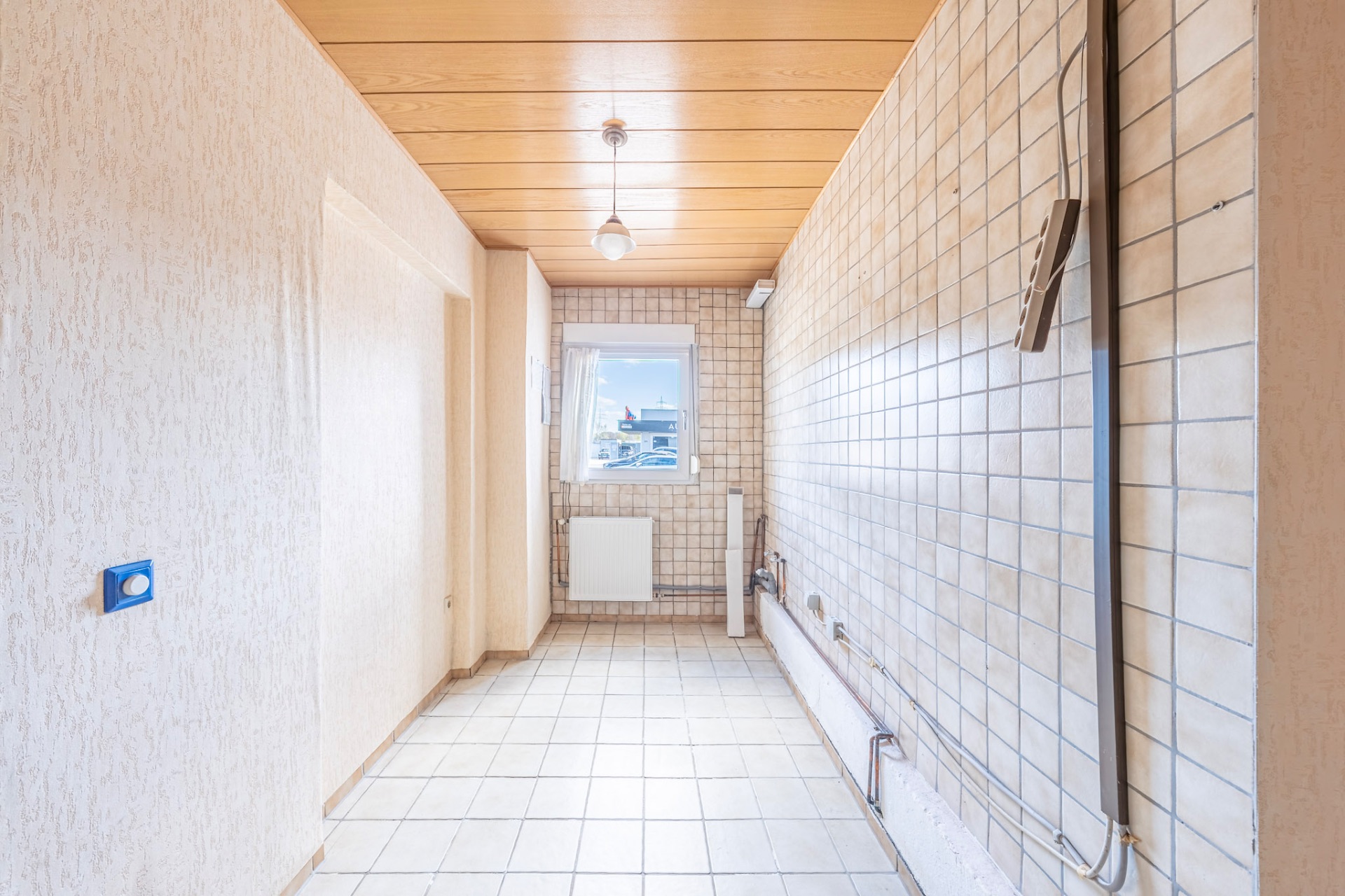 Leerer gefliester Raum mit Fenster, Holzecke und sichtbaren Rohrleitungen an der Wand.