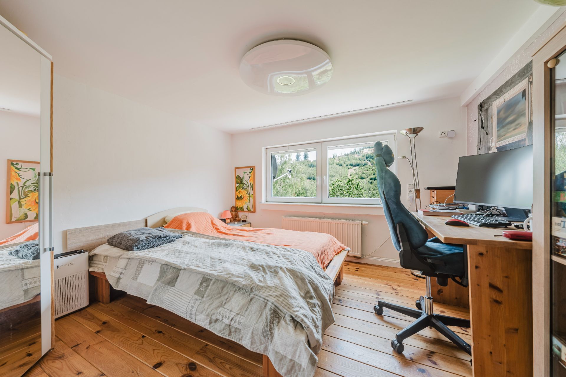 Helles Schlafzimmer mit Holzboden, Bett, Schreibtisch, Stuhl und großem Fenster mit Blick ins Grüne.