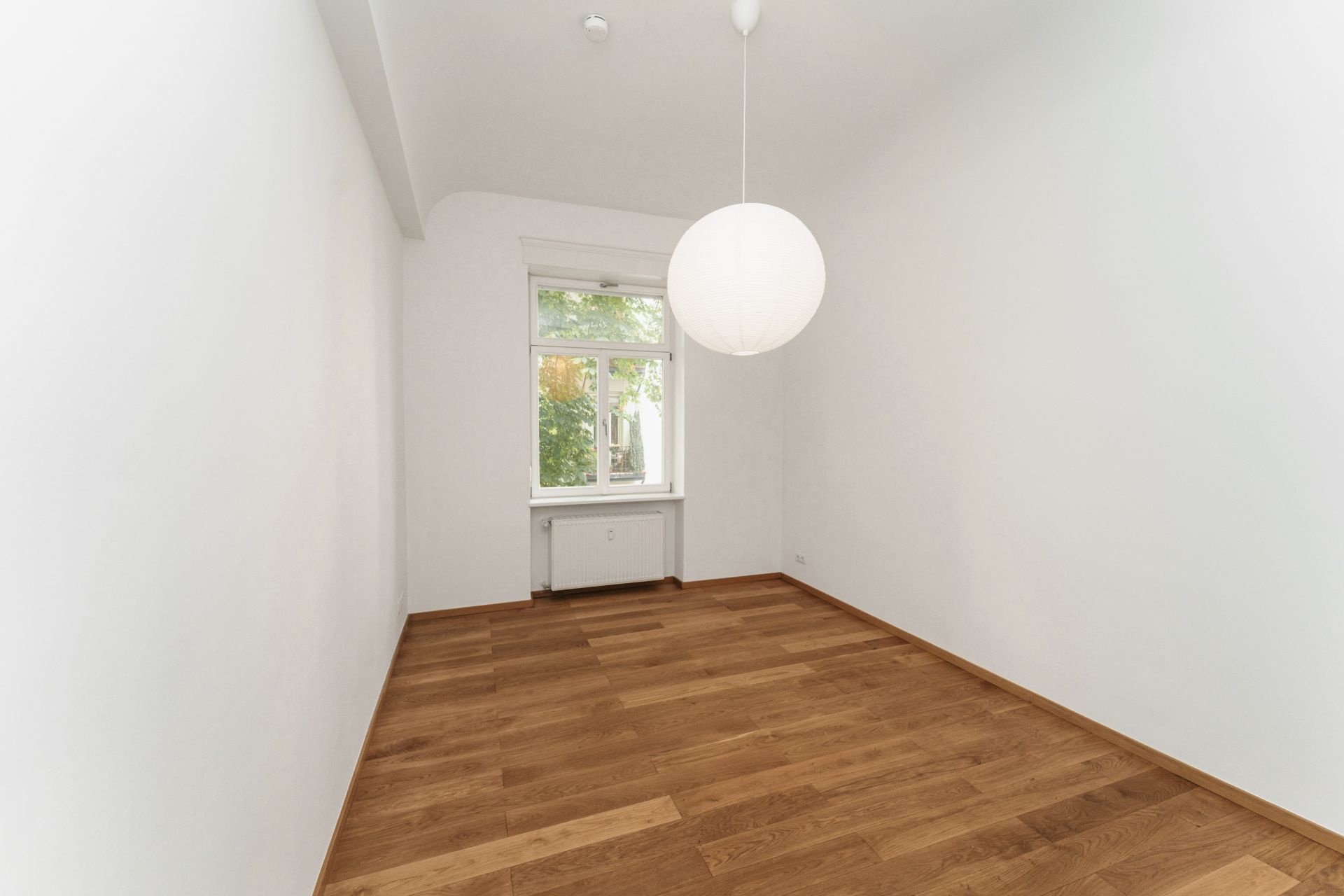 Leerer Raum mit Holzboden, weißer Wand, Fenster und runder Deckenleuchte.