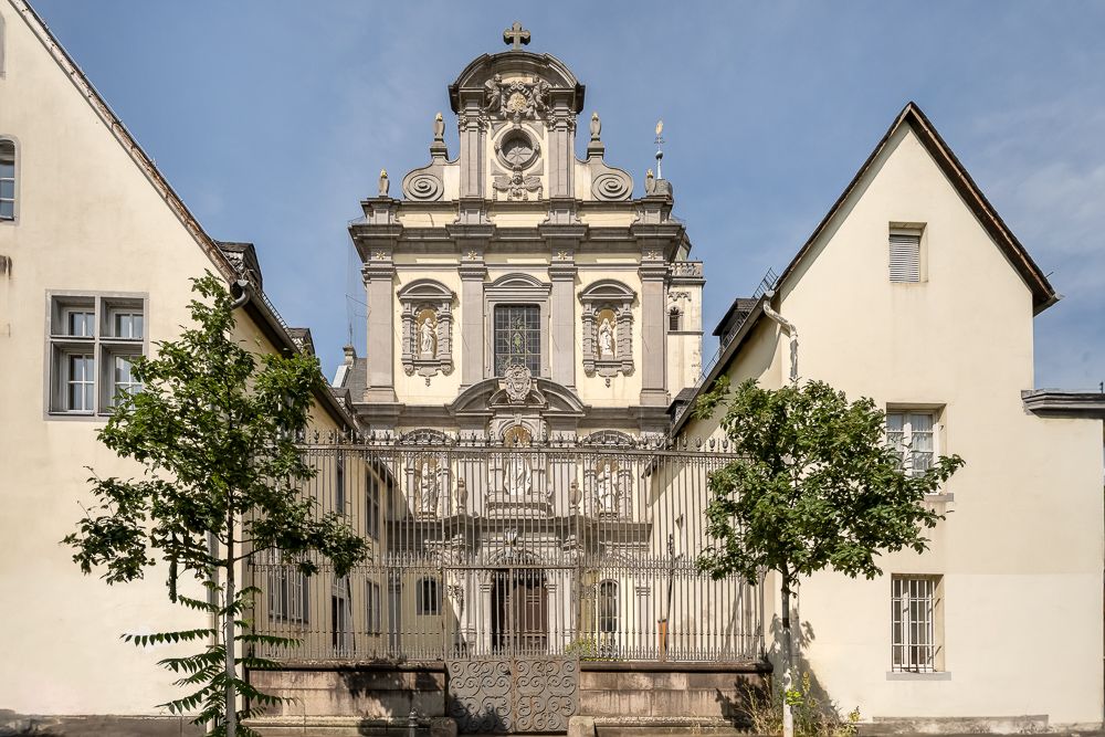 Barockes Gebäude mit schmiedeeisernem Zaun, flankiert von zwei hellen Wohnhäusern und kleinen Bäumen.