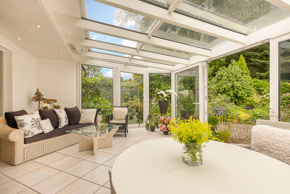 Heller Wintergarten mit Sofa, Glastisch, rundem Tisch und Blick auf grünen Garten.