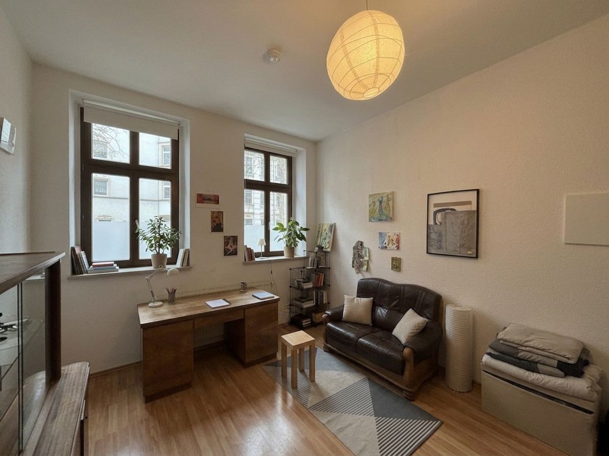 Helles Wohnzimmer mit zwei Fenstern, Schreibtisch, Sofa, Pflanzen und Wandbildern.