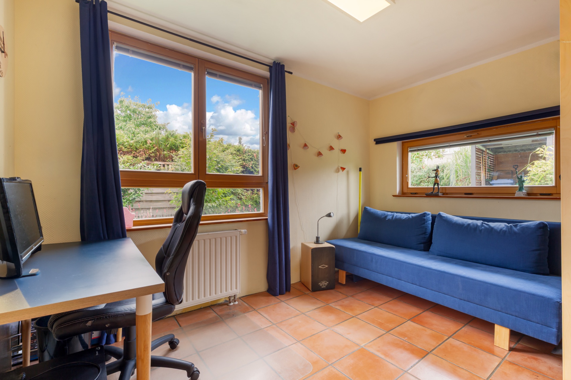 Helles Zimmer mit Schreibtisch, Bürostuhl und blauem Sofa vor großen Fenstern.