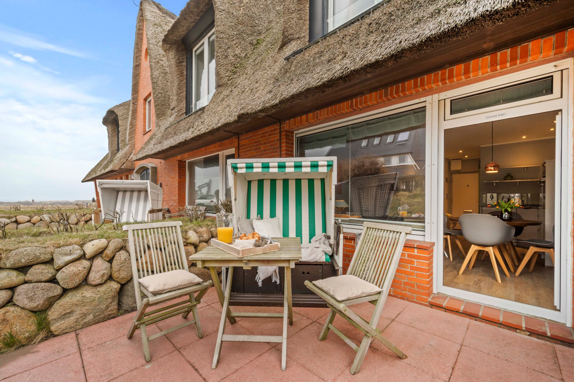 Terrasse mit drei Holzstühlen, einem Tisch und einem Strandkorb vor einem Haus mit Reetdach.