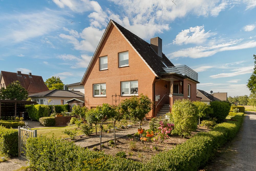 Backsteinwohnhaus mit gepflegtem Vorgarten, Hecken und Blumen unter blauem Himmel.