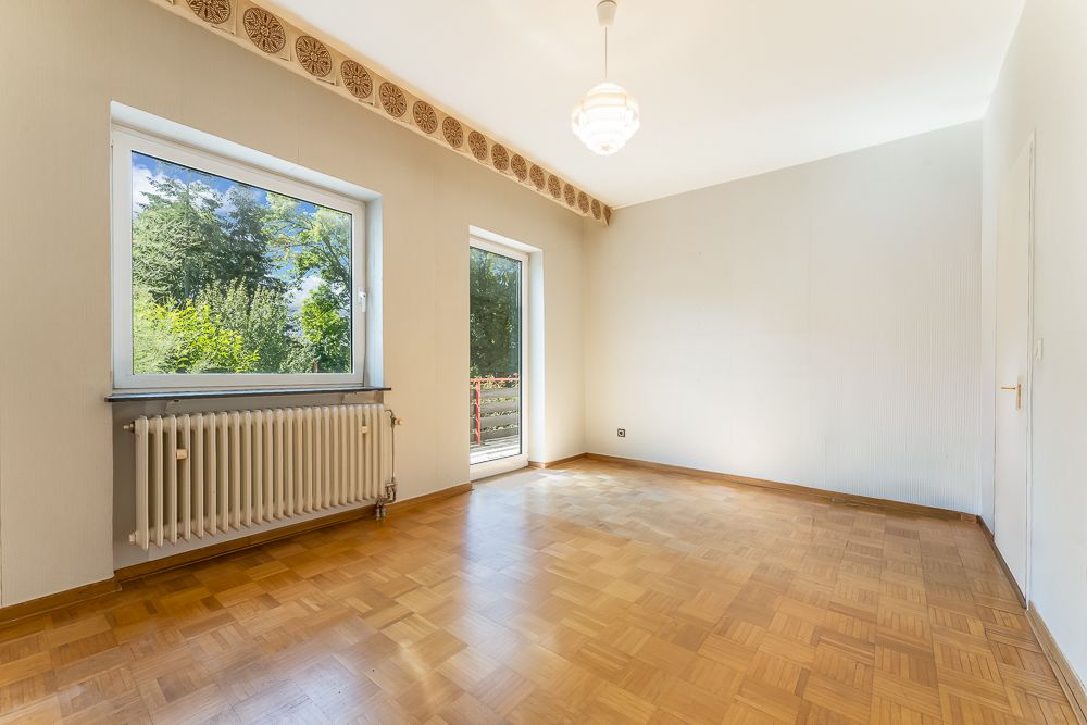 Leerer Raum mit Parkettboden, großem Fenster, Heizung und Zugang zum Balkon.