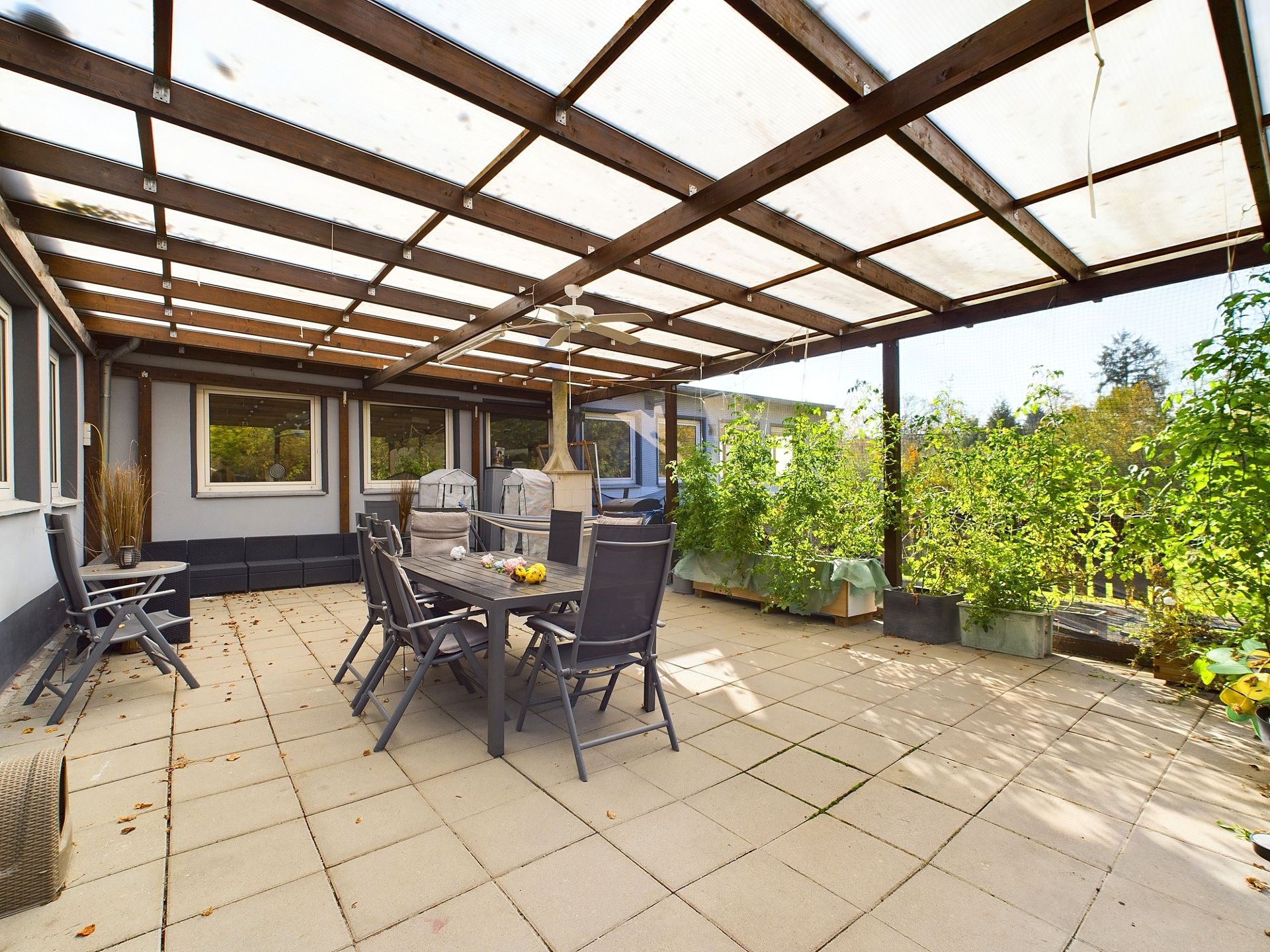 Überdachte Terrasse mit Esstisch, Stühlen und vielen Pflanzen in großen Töpfen.