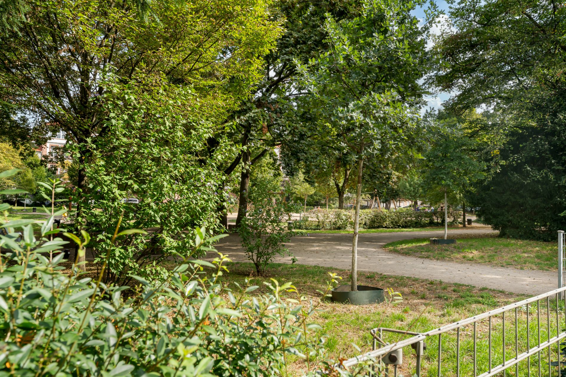 Grüner Garten mit Bäumen, Sträuchern und einem Weg, im Vordergrund ein Metallzaun.