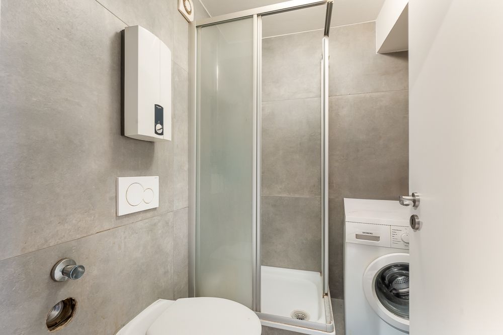 Kleines Badezimmer mit Duschkabine, Waschmaschine und grauen Fliesen an den Wänden.