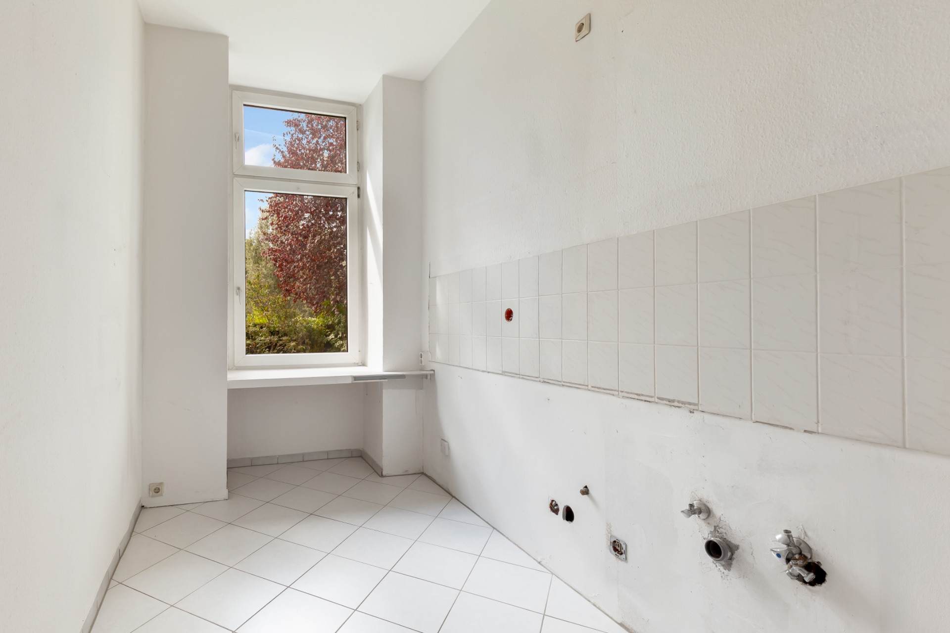 Leerer, gefliester Raum mit großem Fenster und Anschlüssen an der Wand.