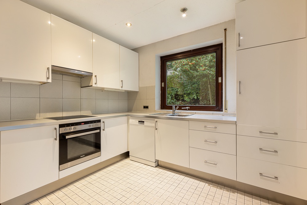 Moderne, helle Küche mit weißen Schränken, Herd, Spülmaschine und Fenster mit Blick ins Grüne.
