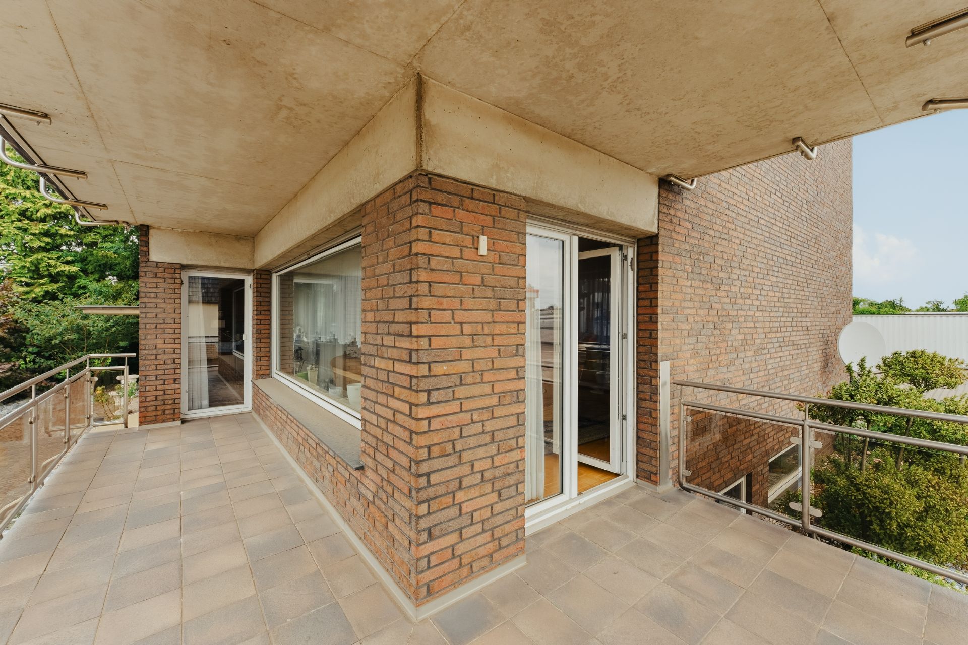 Großer Balkon mit Fliesenboden, Ziegelwand und Glasschiebetür, umgeben von Metallgeländer.