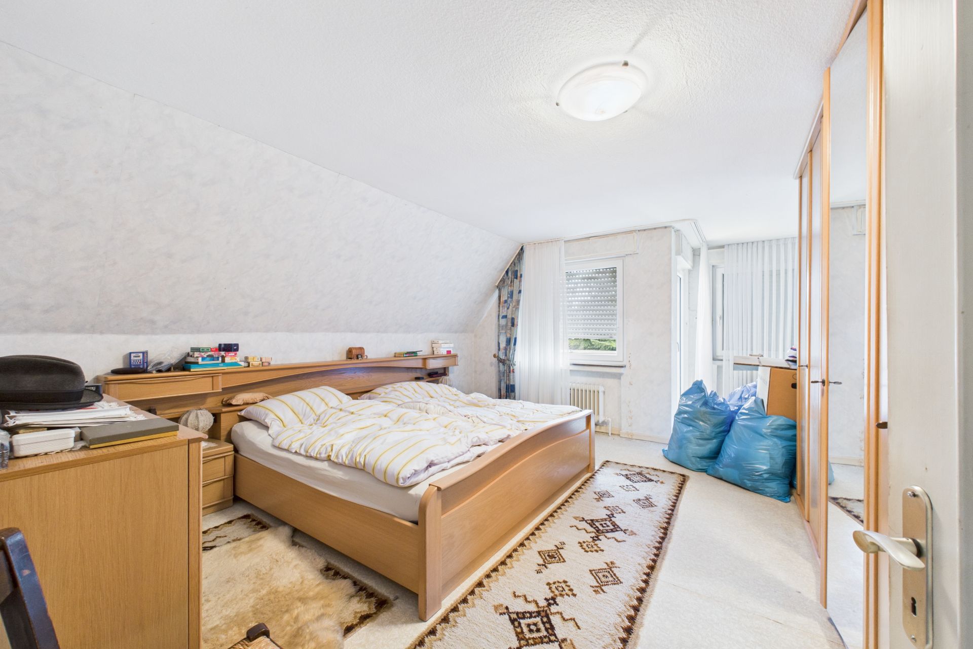 Helles Schlafzimmer mit Doppelbett, Kleiderschrank mit Spiegel und zwei blauen Säcken am Fenster.