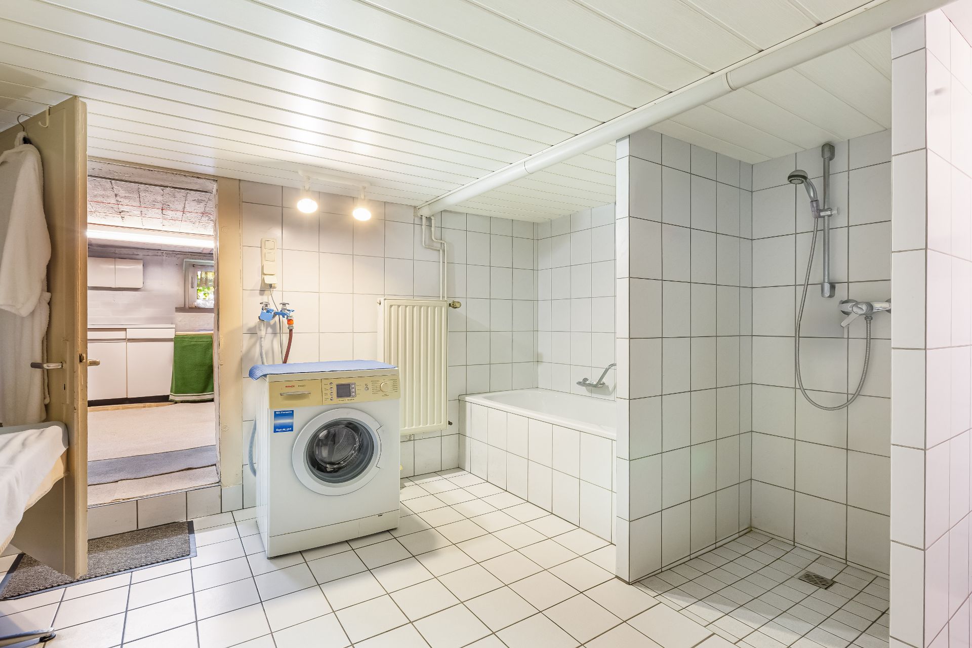 Helles Badezimmer mit Dusche, Badewanne und Waschmaschine auf weißen Fliesen.