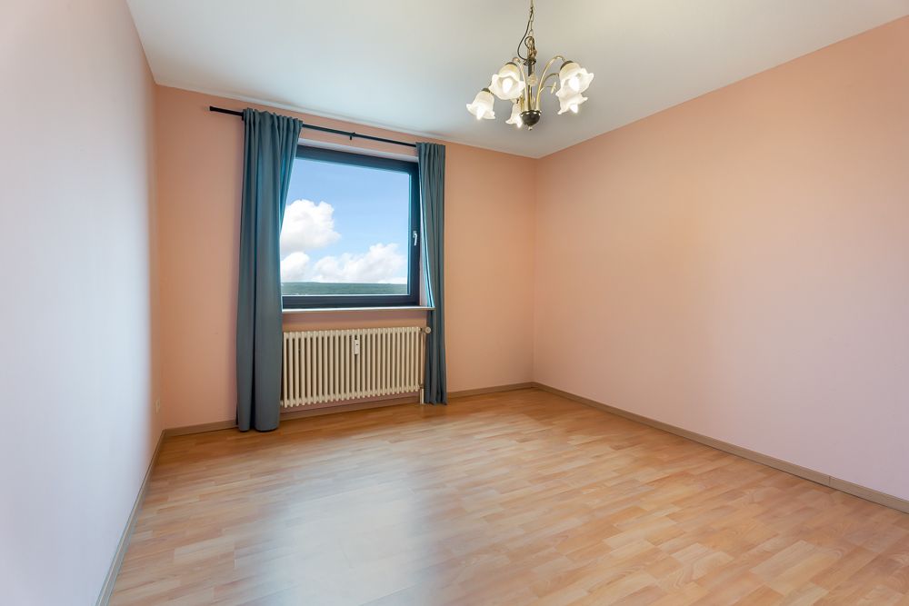 Leeres Zimmer mit hellen Wänden, Laminatboden, Fenster mit Vorhängen und Deckenleuchte.