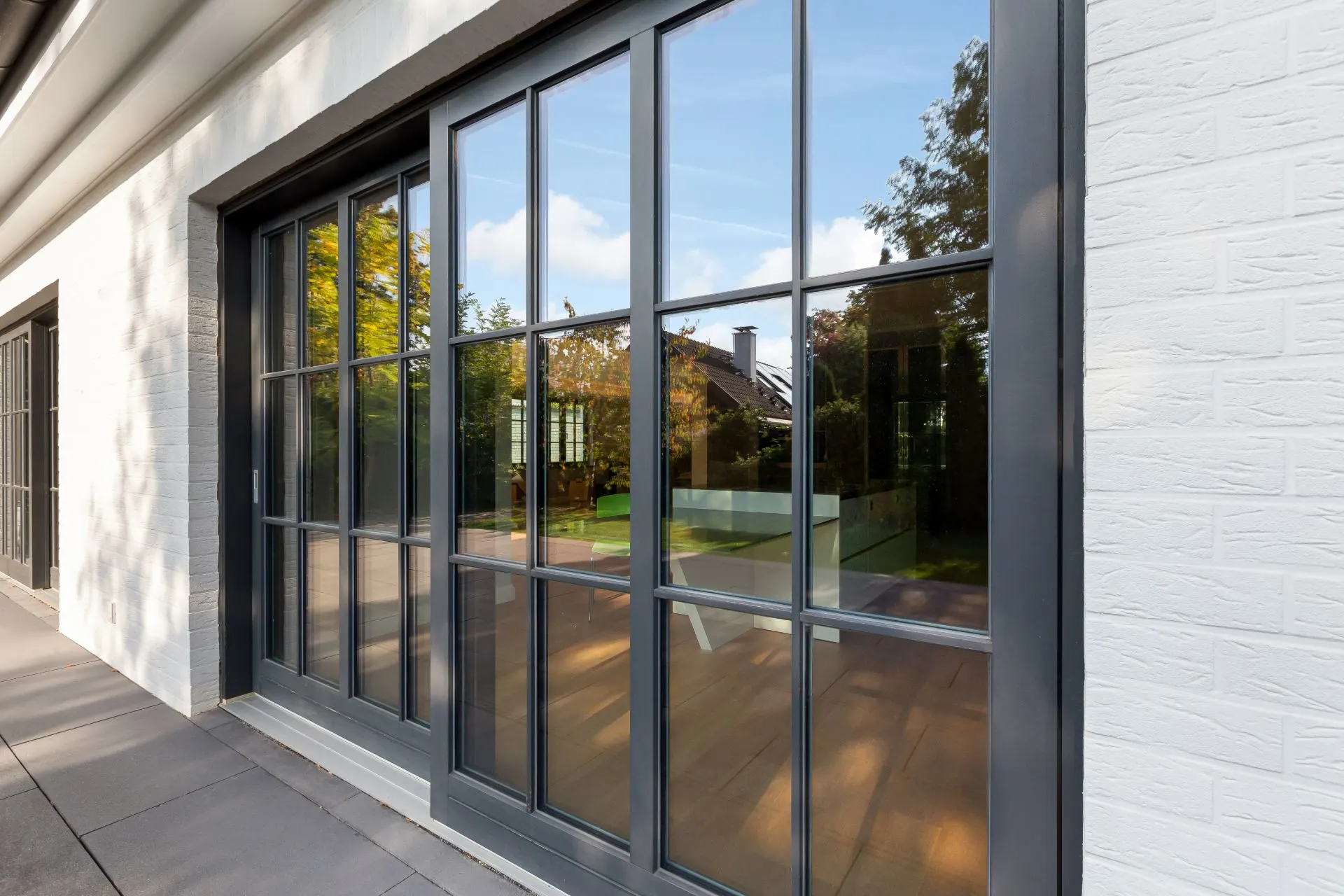 Große Schiebetür aus Glas mit schwarzem Rahmen an einem Wohnhaus, Spiegelung von Bäumen sichtbar.