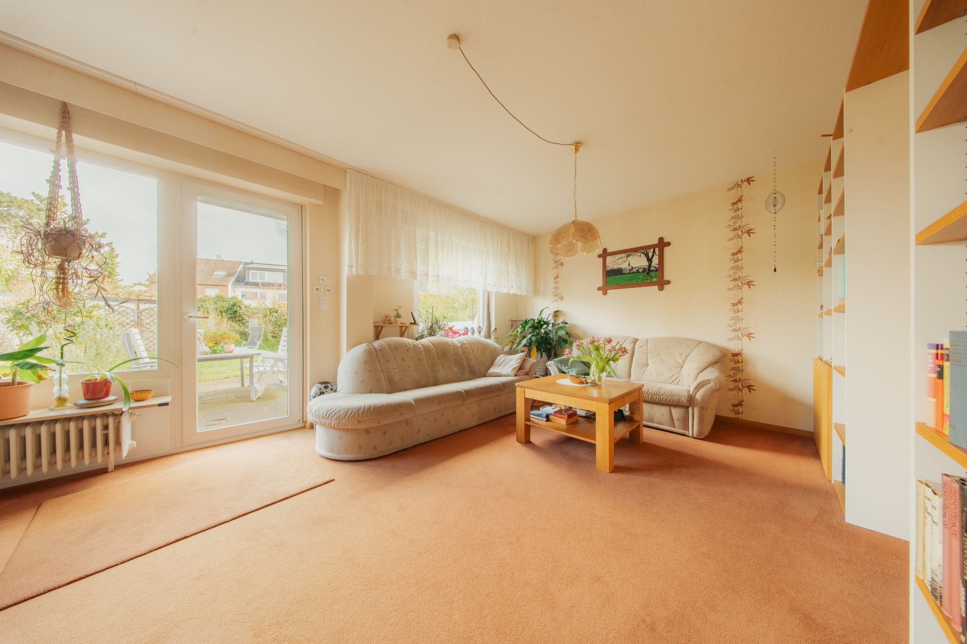 Helles Wohnzimmer mit Sofa, Couchtisch, Bücherregal, Pflanzen und großen Fenstern zum Garten.