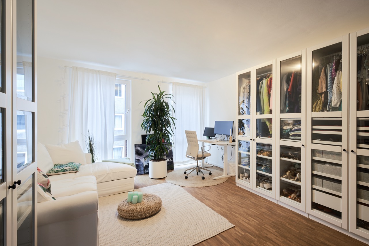 Helles Wohnzimmer mit Sofa, Schreibtisch, Pflanze und großer Schrankwand mit Glastüren.