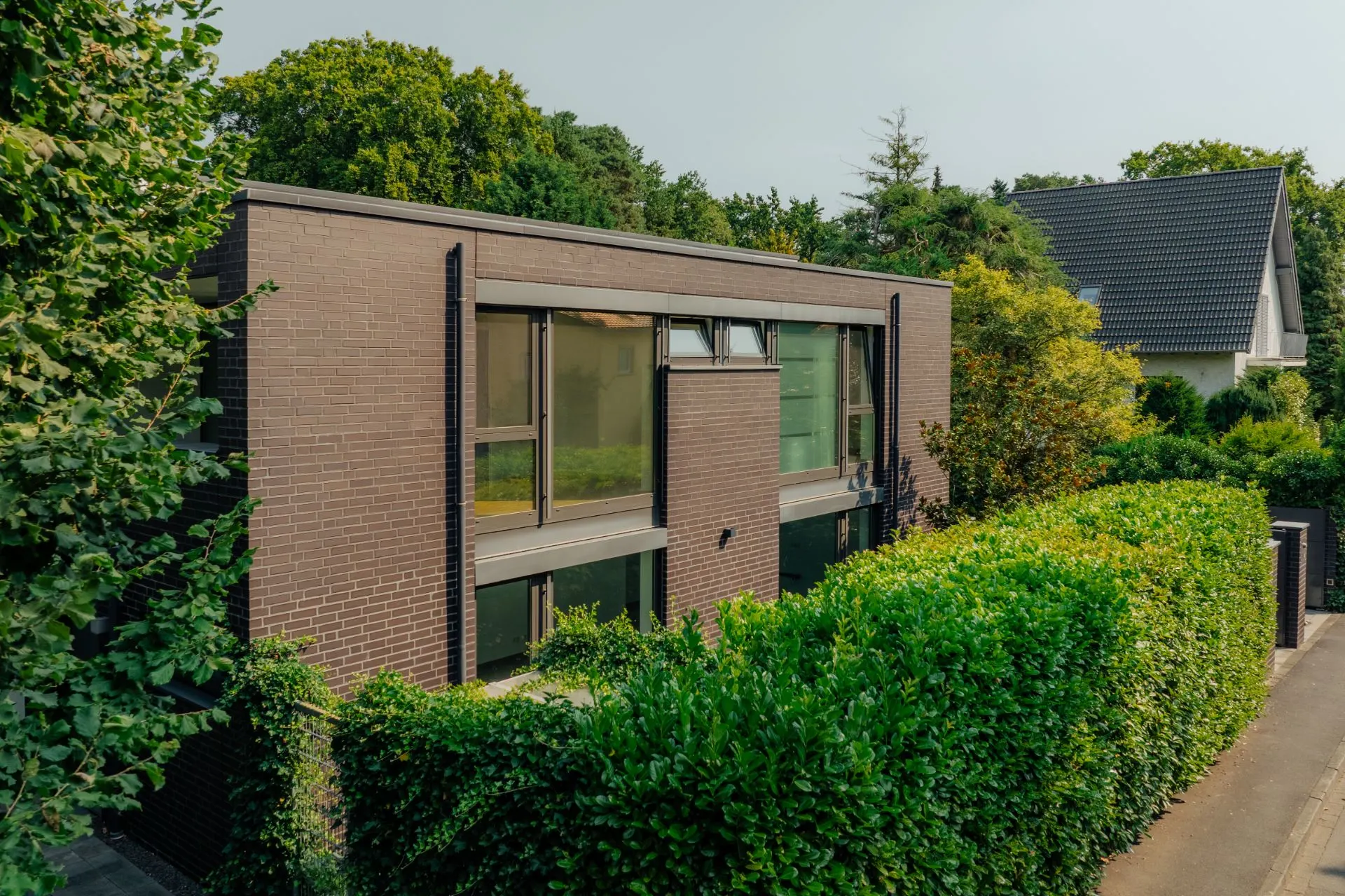 Modernes zweistöckiges Haus mit großen Fenstern und Backsteinfassade, umgeben von grüner Hecke.