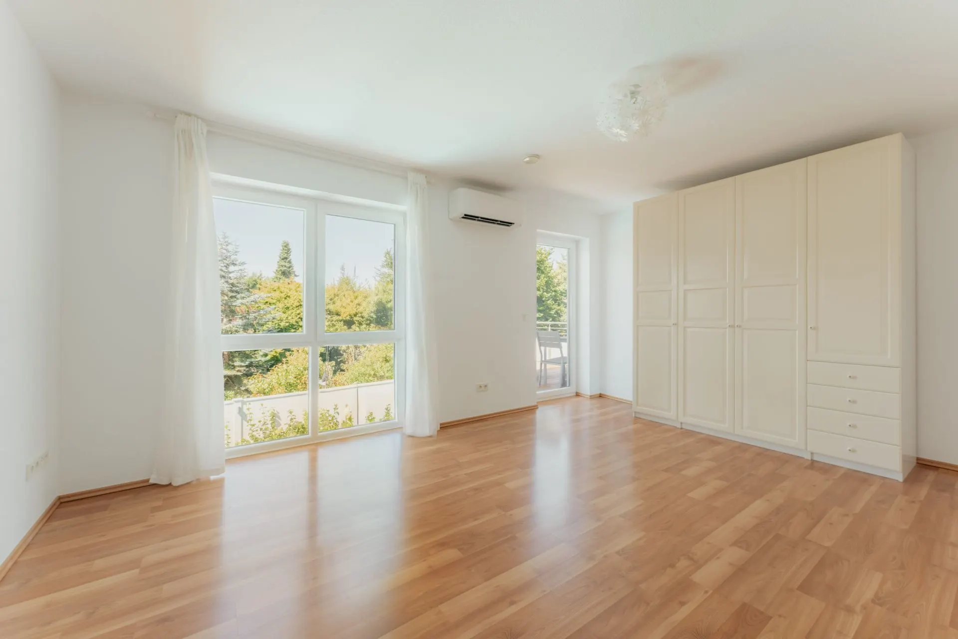 Helles Zimmer mit Holzfußboden, großen Fenstern, Gardinen und weißem Einbauschrank.