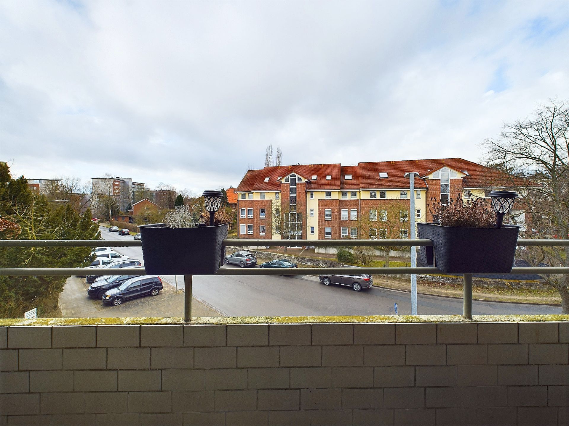 Blick von einem Balkon auf eine Straße mit geparkten Autos und ein gegenüberliegendes Wohnhaus.