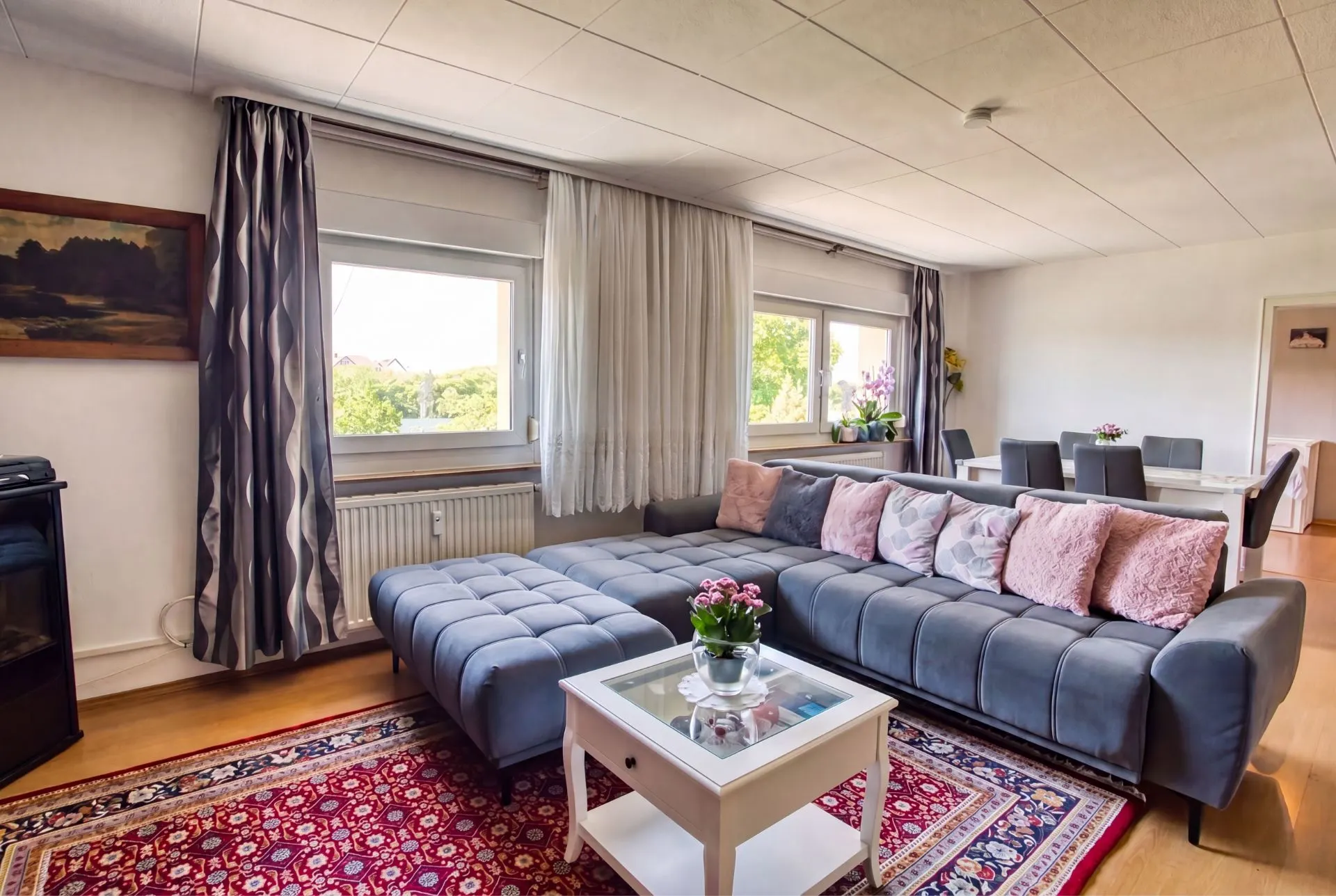 Helles Wohnzimmer mit grauem Sofa, Teppich, Couchtisch und Essbereich im Hintergrund.