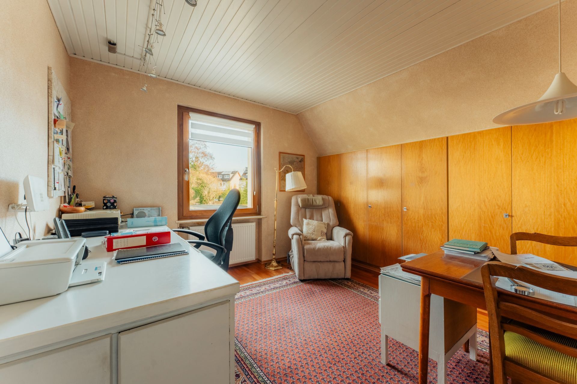 Helles Arbeitszimmer mit Schreibtisch, Bürostuhl, Sessel, Teppich und großen Holzschränken.