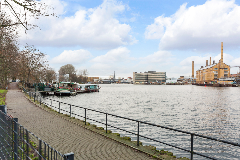 Gepflasterter Weg mit Geländer entlang eines Flusses, Boote und Gebäude im Hintergrund.