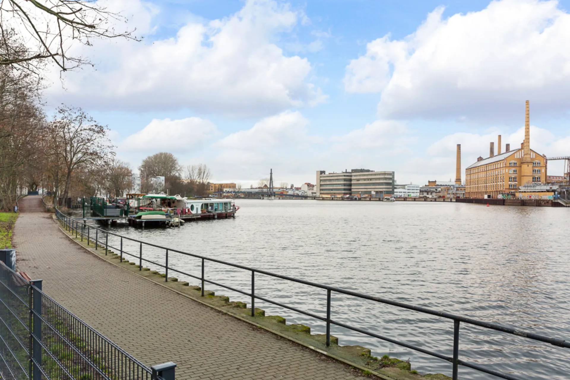 Gepflasterter Weg mit Geländer entlang eines Flusses, Boote und Gebäude im Hintergrund.