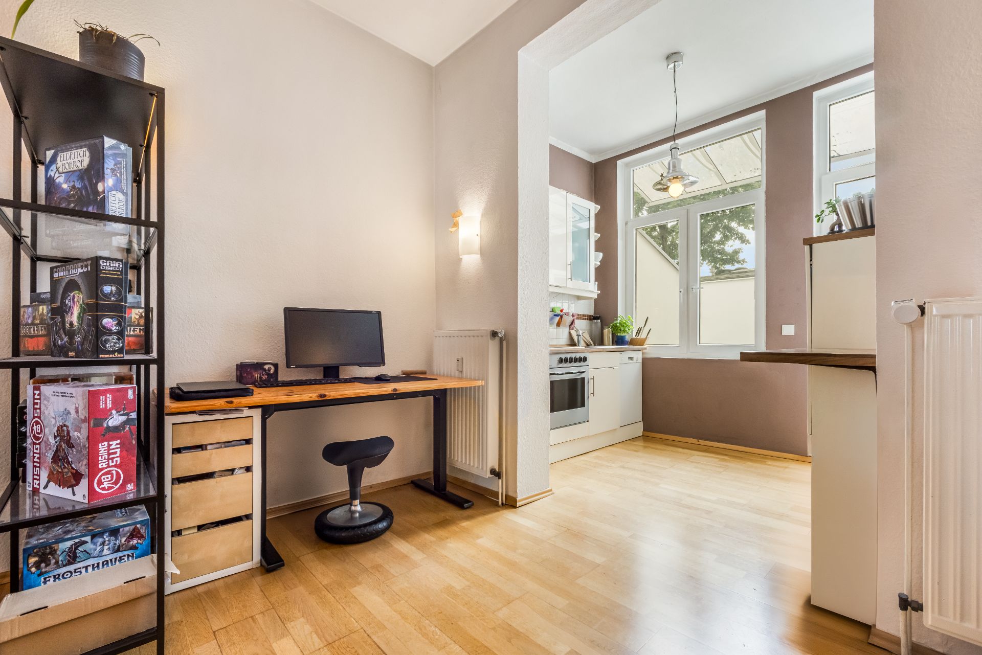Arbeitsbereich mit Schreibtisch und Computer neben offener Küche mit großen Fenstern.